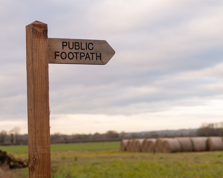 A wooden sign reading "Public Footpath" is photographed over a green pasture with a treeline in the background in a cloudy, overcast sky.