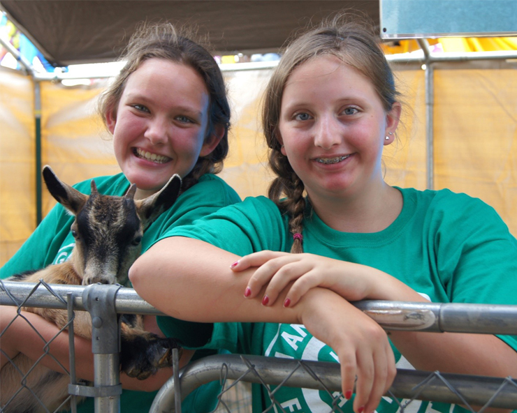 Closeup of 2 girls in green T-shirts, one holding a goat and the other leaning on a fence